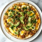 Top-down view of a freshly baked breakfast pizza with eggs, cheese, and seasoned chicken on a white plate.
